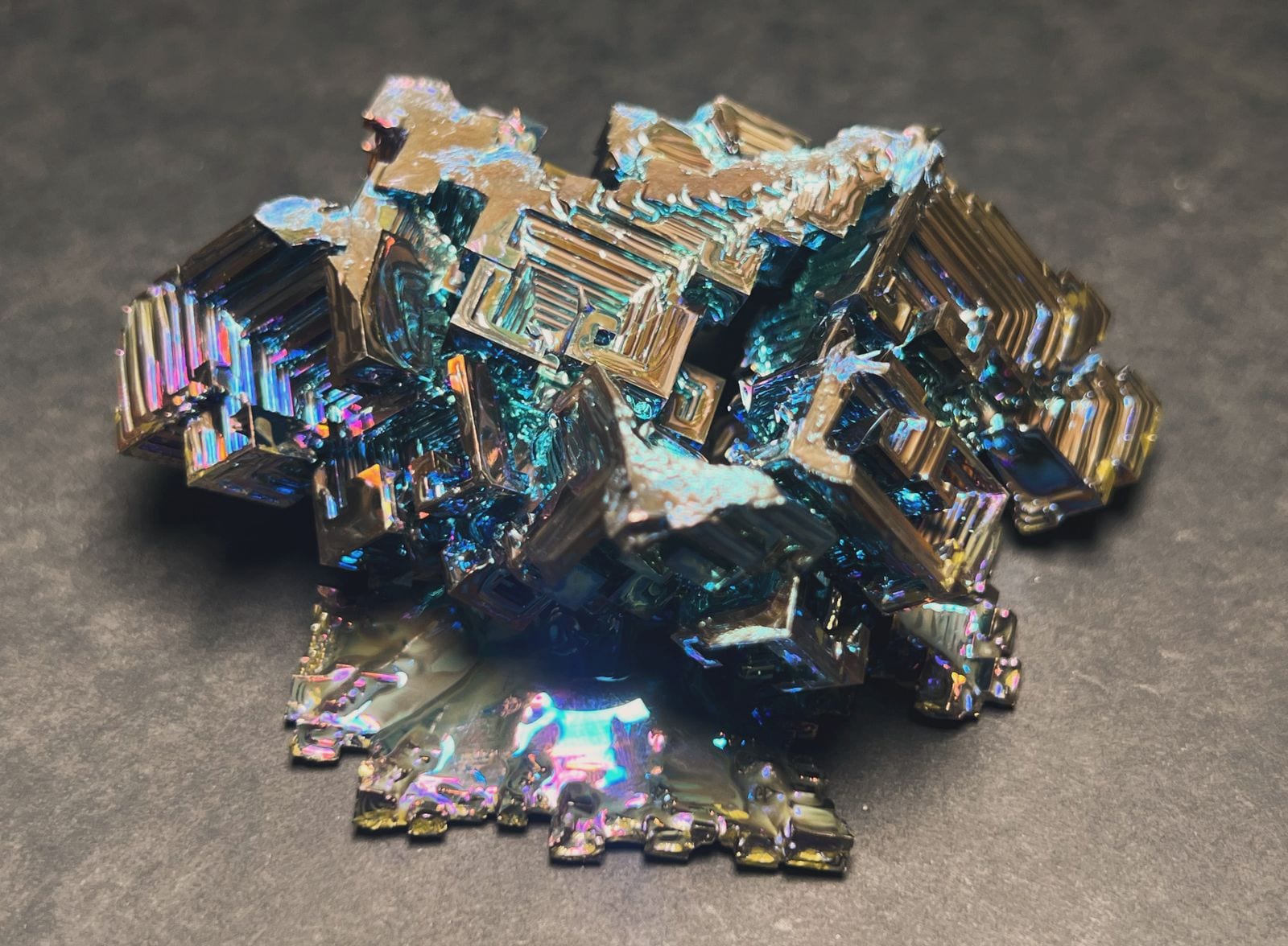 Iridescent rainbow bismuth crystal with stair-stepped hopper geometry catching blue, gold, and violet light on a slate-gray surface